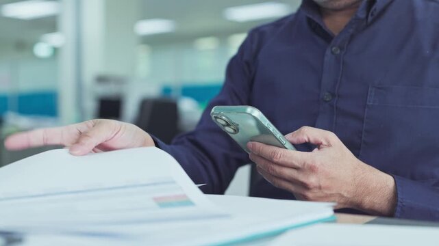 Businessman holding mobile phone examines corporate report sheets at office desk, reviewing marketing paperwork cross referencing financial data during daytime corporate workflow.