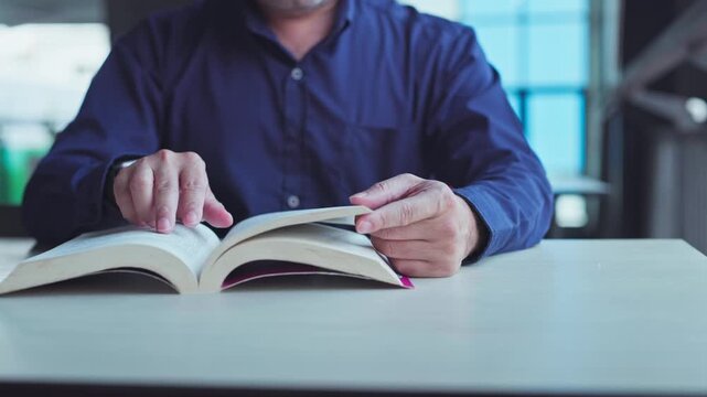Mature man sitting indoor desk turning pages open textbook studying quiet academic room daylight.