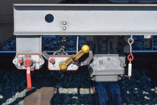 A close up industrial shot shows the mechanical brake valve changover levers with yellow and red handles mounted under a train freight car chassis on a sunny day.