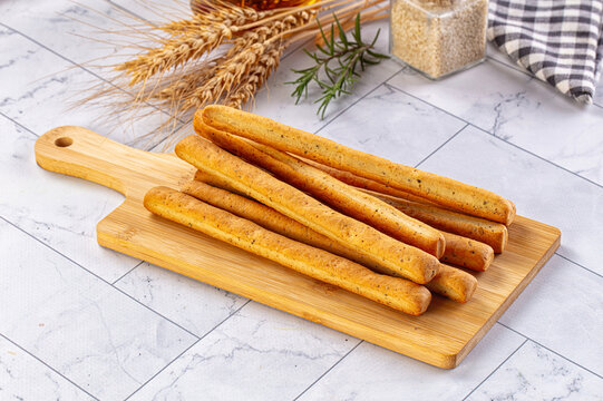 Crispy grissini breadsticks piled on wooden serving board with wheat ears, rosemary sprig and sesame seeds on marble kitchen table, Italian snack food
