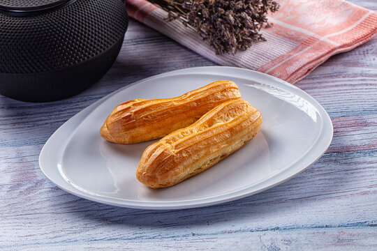White ceramic plate holding two golden brown eclair pastries beside black textured speaker and dried lavender on striped napkin, dessert presentation