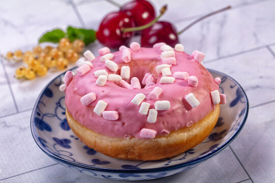 Pink glazed donut topped with mini marshmallows on blue patterned ceramic plate with fresh cherries and white currants in background, sweet pastry dessert