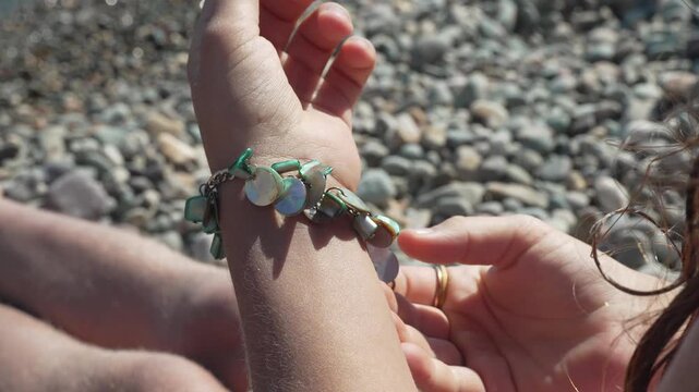 Decorative beaded seashell jewelry fastened by young person sitting near coastal pebble shore. Close up tracking shot.