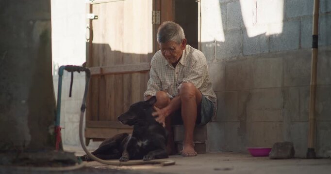 A man who is senior sits on his doorstep petting a black dog, barefoot in the warm evening sunlight of a weathered old house.
