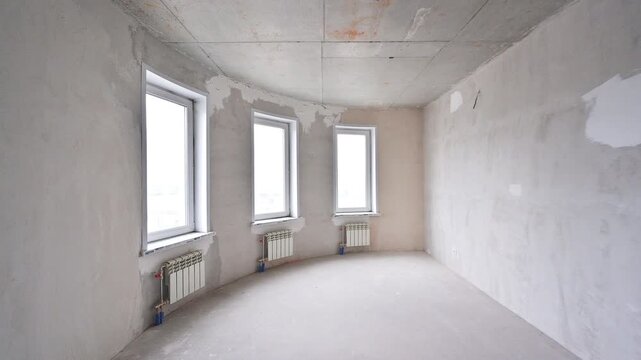 Sparse room in pre‑finish stage. three windows with white frames, radiators below, rough plaster walls, and exposed ceiling edges
