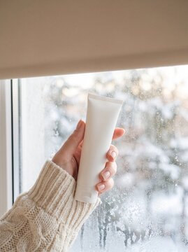 Hand in knit sweater holding blank hand cream tube by window