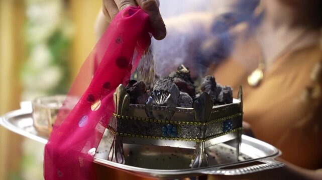 Detailed close up capturing the aromatic smoke rising from wild rue burning on hot red charcoal inside a traditional metal brazier used for spiritual protection at a cultural wedding ritual.
