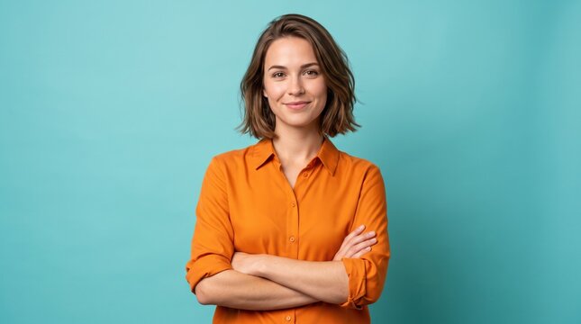Confident young woman standing with arms crossed wearing an orange buttoned shirt against a solid turquoise background looking directly at the camera with a friendly and positive expression