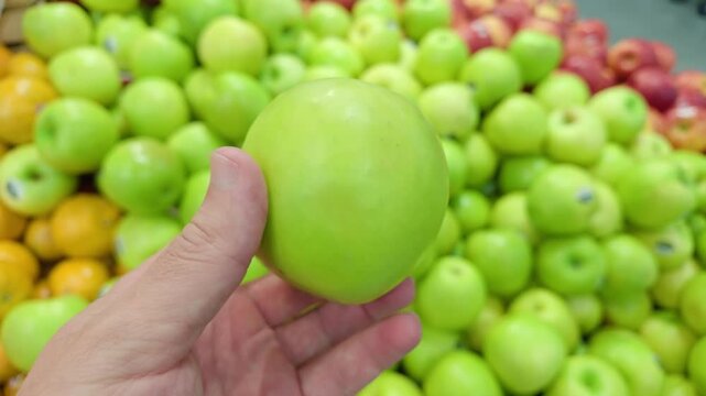 Close-up of a man holding and choosing a fresh green apple from a large display at a supermarket or grocery store. Retail trade, fresh produce, food shopping and healthy nutrition concept.