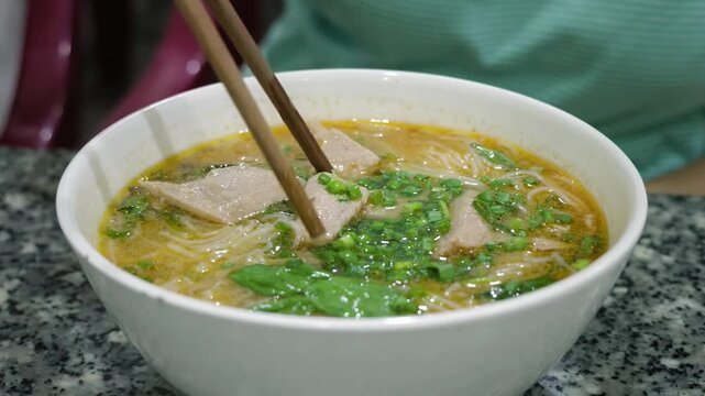 Close-up of a person using chopsticks to eat traditional Vietnamese noodle soup with fish, herbs and broth in a white bowl. Asian street food, local cuisine and authentic dining concept.