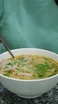 Close-up of a person using chopsticks to eat traditional Vietnamese noodle soup with fish, herbs and broth in a white bowl. Asian street food, local cuisine and authentic dining concept.