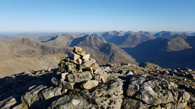 A stone cairn marks the rocky summit of a mountain with a vast panoramic view of distant rugged peaks under a blue sky