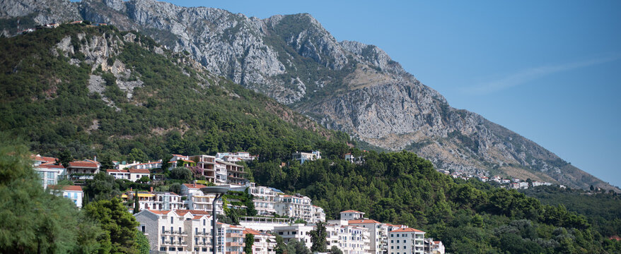 Cluster of white-walled, red-roofed buildings climbs up a steep, lush green hillside beneath a towering, rocky mountain