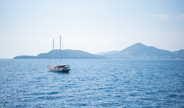 Two-masted wooden sailboat cruises on the calm blue sea near a mountainous coastline