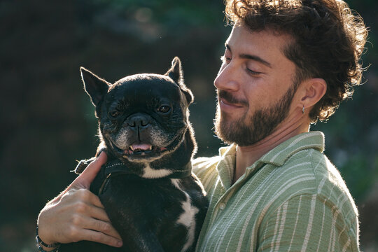 Young man smiling while holding a French bulldog outdoors in natural sunlight