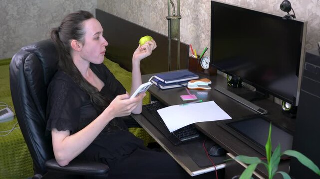 A woman sits at a desk in a home office. She eats an apple while looking at her phone. Papers and a computer are on the desk, showing a busy workspace.