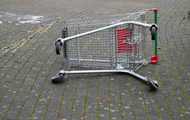 An overturned metal shopping cart lies abandoned on its side on a grey cobblestone street, symbolizing vandalism, neglect, or urban decay. © petra goeschel