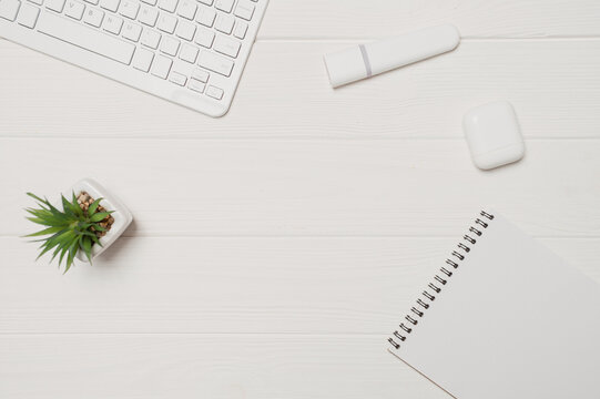White minimal office workspace flat lay with modern wireless keyboard earbuds and notepad clean wooden desk