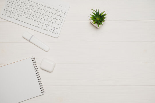 White minimal office workspace flat lay with modern wireless keyboard earbuds and notepad on clean wooden desk