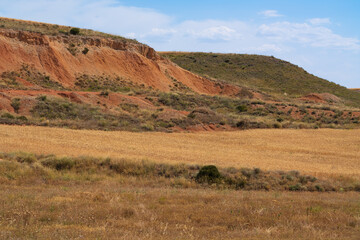 An eroded red clay hillside with exposed orange soil layers rises above a golden harvested wheat field and sparse green shrubs. White clouds drift across a bright blue sky over this dry rural valley © Aimur