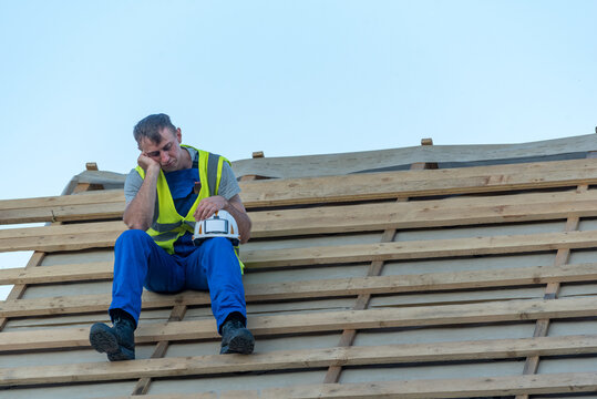 Exhausted construction worker in blue uniform and safety vest rests on wooden roof battens, holding his hard hat while taking a break under a clear sky after long hours