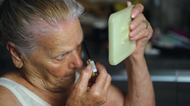 Elderly woman aged 87 carefully applying mascara to her eyebrows and eyelashes in her kitchen studio while maintaining her personal appearance