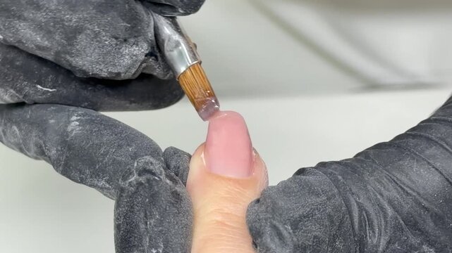 Macro close‑up of a manicure . A nail technician in black gloves applies a thick nude pink camouflage builder gel with a brush to level and reinforce the big toenail