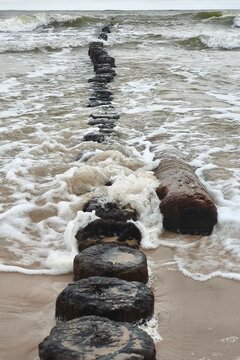 ​Old wooden breakwater poles stretching into foaming sea waves on a sandy beach shore, capturing coastal erosion defense, dramatic marine landscape, and outdoor nature.