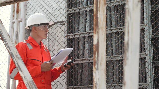 Engineer checks technical documents beside fenced electrical transformer, recording inspection findings during industrial facility survey. Power distribution equipment, maintenance assessment.