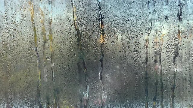 Water droplets running down foggy window glass, creating condensation streaks. Cold outside weather causes moisture buildup on glass surface, making it difficult to see through. Blurry view indoor