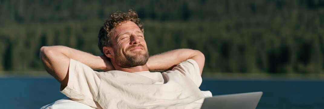 Bearded man relaxing on a lake shore with a laptop for remote work wallpaper and serene outdoor leisure background