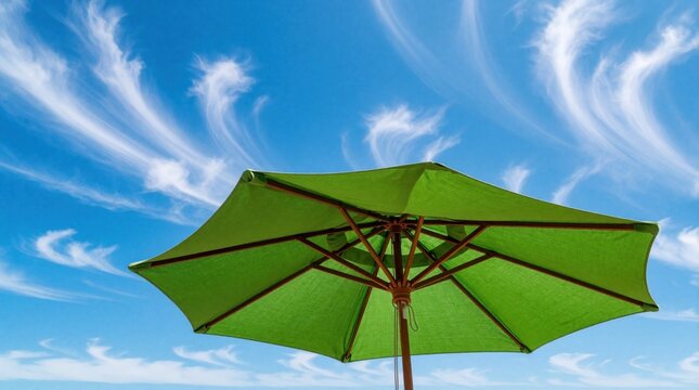 Green garden parasol stands under bright blue sky with wispy white clouds. Large lime sun umbrella provides shade in summer yard. Outdoor patio furniture offers shelter from hot sun, heat during