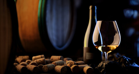 Naklejka premium Golden white wine in elegant glass on dark background. Wine cellar of vinery with barrel and corks.