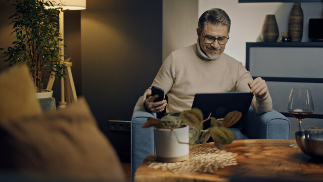Middle aged man sitting in comfort in living room working on laptop computer and phone in home office. Businessman managing business online. Portrait of confident older male, happy, smiling.