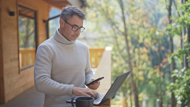 Mature man with smartphone and laptop working remotely on terrace of wooden chalet in forest. Serenity and peaceful relaxed lifestyle. Businessman using digital technology and telecommunication.
