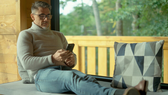 Relaxed mature man using smartphone in cozy wooden cabin, enjoying peaceful moment by window with scenic forest view. Mid adult male in 50s in turtleneck pullover and glasses reading on phone.