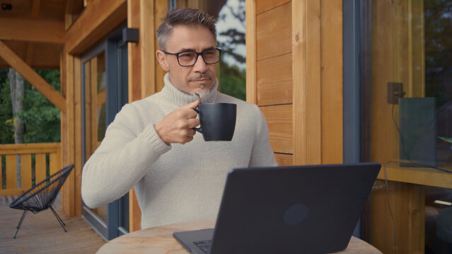 Mature man drinking hot coffee working remotely on laptop computer, outdoor on porch of mountain chalet. Businessman in home office. Entrepreneur remote managing business from weekend house.