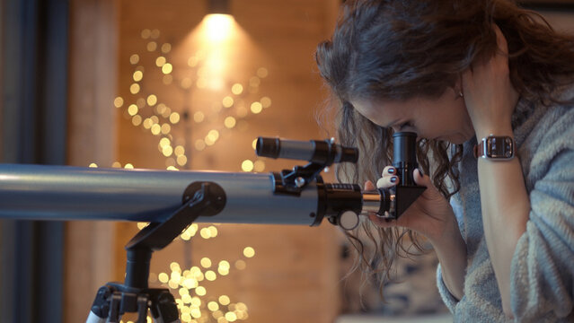 Middle aged woman at home looking out window with telescope. Female in turtleneck pullover standing in living room of wooden cabin with Christmas lights. Autumn or winter indoor clothing.