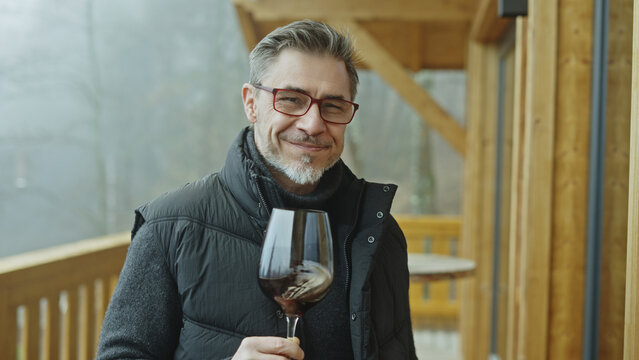 Expert drinking, tasting glass of red wine. Middle aged man outdoor on home balcony wearing turtleneck pullover and warm vest. Eyeglasses and gray hair. Happy satisfied smiling. Autumn or winter.