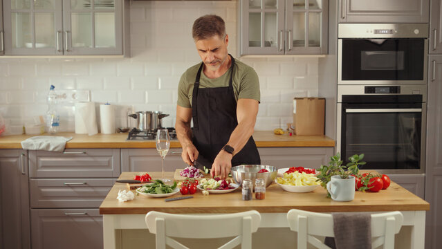 Mid adult man preparing for cooking in modern kitchen at home, wearing casual cloth and apron. Male chef following food recipe, cleaning and slicing vegetables.
