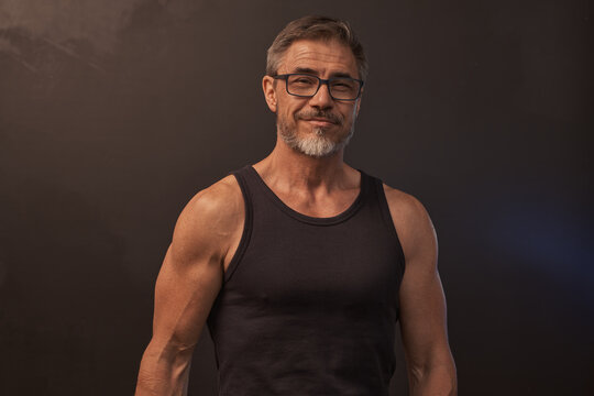 Portrait of happy muscular athletic mid adult man standing against dark background, smiling. Studio shot of middle aged confident sporty male fitness trainer with gray hair in eyeglasses.