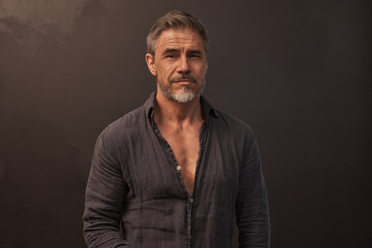 Portrait of mid adult man standing against dark background in gray casual clothes. Studio shot of middle aged confident sporty male with gray hair.