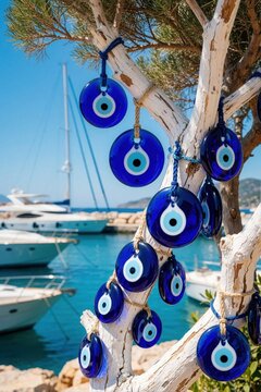 Blue Turkish evil eye charms hang on white tree branch. Nazar amulet ornaments provide protection, luck. Sunny summer day at mediterranean marina with white yachts, turquoise sea background.