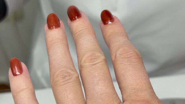  Macro close‑up of a salon manicure. A nail technician in black gloves applies a burgundy/brown‑red gel polish with a bottle brush: the pinky is painted first, then the thumb. Visible prep dust on the