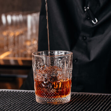 Amber beverage poured into a cut glass tumbler over a textured bar mat with bartender in black clothing, close up of a bar counter