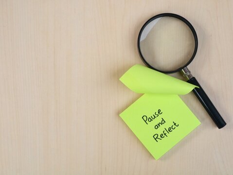 Pause and reflect handwritten text on green adhesive note with magnifying glass on wooden desk background. Self evaluation, mindfulness, review, and business strategy concept with copy space