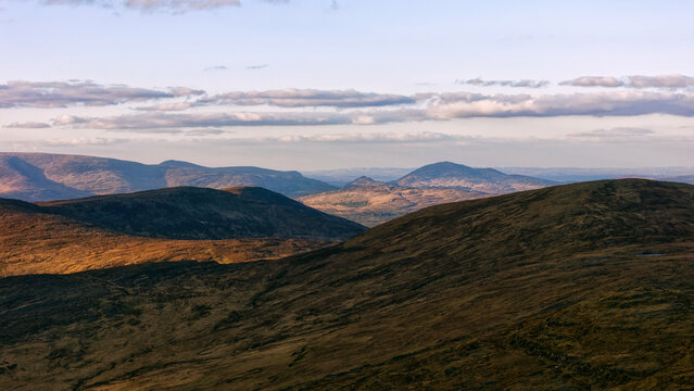 Golden afternoon light illuminates the vast grassy slopes and distant mountain ranges in Ireland as soft clouds drift across the horizon during the daytime.