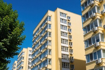Fototapeta premium Modern yellow apartment buildings under clear blue sky in summer. Multi story residential blocks with many windows, air conditioners on facade. Urban housing estate with green trees in city district.