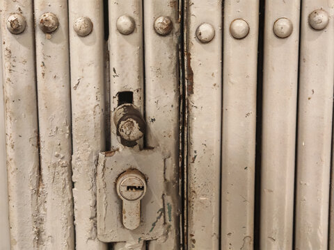 Extreme close-up of a heavy-duty grey painted metal folding security gate. Features a central lock cylinder, handle, mechanical rivets, vertical structural lines, specular highlights