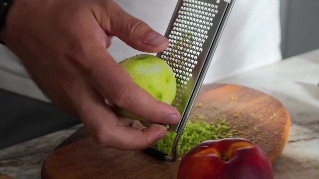 Close-up shot of hands zesting lime with grater over wooden cutting board, with ripe peach nearby, preparing ingredients for cooking or baking.